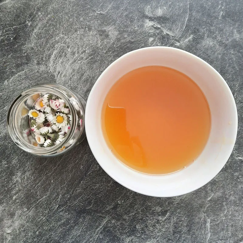 Glas mit Blüten des Gänseblumchens neben Schale mit Essig-Honig-Mischung Zur Herstellung des Gänseblümchenoxymels